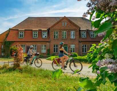 Zwei Frauen auf ihren Fahrrädern fahren am Gutshaus Linstow vorbei, sie schauen zum Gutshaus