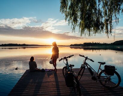 Zwei Frauen befinden sich auf einem Steg der ins Wasser ragt. Eine der Frauen sitzt und blickt aufs Wasser, die andere steht hinter ihr und hält einen Fahrradhelm in der Hand. Langsam geht die Sonne unter und hinter ihnen stehen zwei Fahrräder.