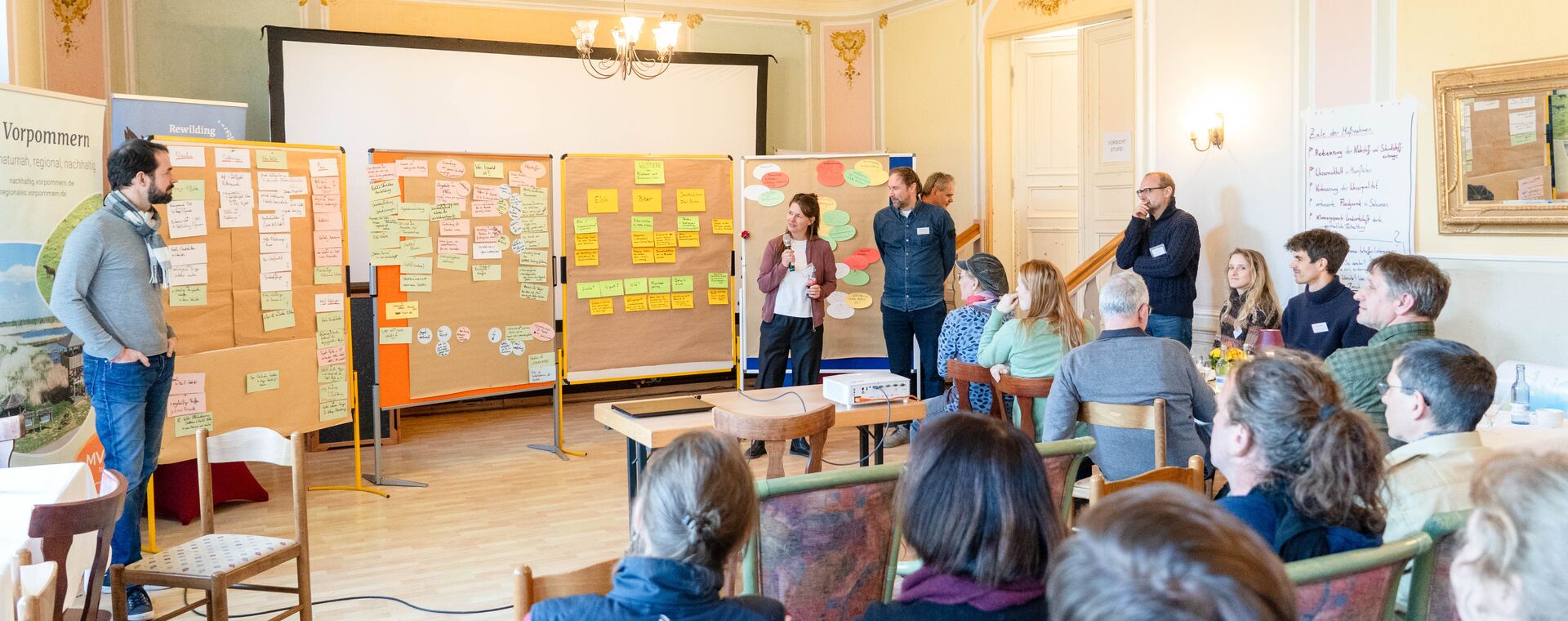 Eine Gruppe an Menschen hört aufmerksam zwei Moderatoren zu, die die Ergebnisse eines Workshops präsentieren
