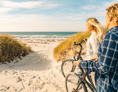 Auf dem Bild sieht man den Blick aufs Meer auf der Insel Hiddensee durch die Dünen. Rechts im Bild stehen ein Mann und eine Frau mit dem Rücken zur Kamera, die ihre Fahrräder Richtung Meer schieben.