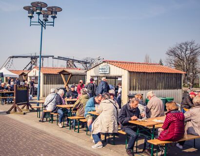 Viele Menschen auf Bierbänken und an Tischen am Essen. Neben den alten Bootshütten am Hafen in Wieck