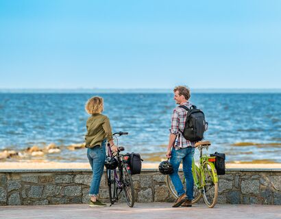 Ein Paar steht mit seinen Fahrrädern vor einer kleinen Steinmauer und blickt aufs Meer. Neben ihnen ist ein Fahnenmast und daran hängt eine gelbe Fahne