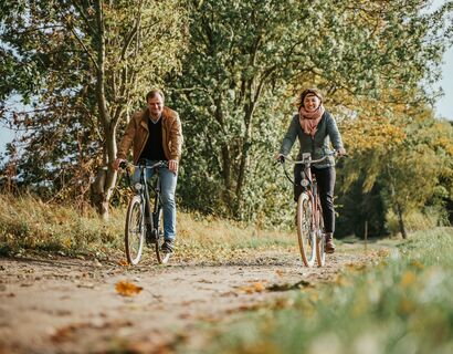 Ein Mann und eine Frau fahren auf Fahrrädern über einen Feldweg. Auf dem Boden liegen Blätter und die Bäume sind schon leicht herbstlich gefärbt