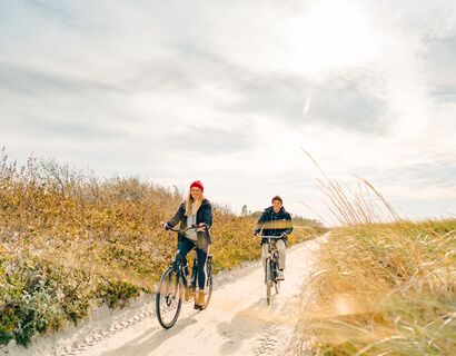Ein Mann und eine Frau fahren auf Fahrrädern über einen Sandweg durch die Dünen. Der Weg ist umgeben von Sträuchern und Strandhafer