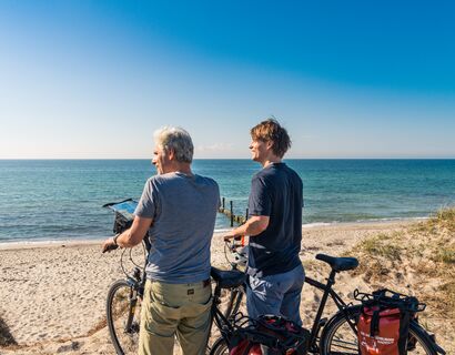 Zwei Männer stehen mit dem Rücken zur Kamera und zum Meer gerichtet am Strand mit ihren Fahrrädern und blicken auf das türkise Wasser. Sie sehen zufrieden aus