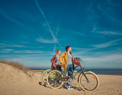 Am Strand sind zwei Frauen, die ihre Fahrräder durch den Sand schieben. Links ist eine kleine Düne und im Hintergrund das Wasser.