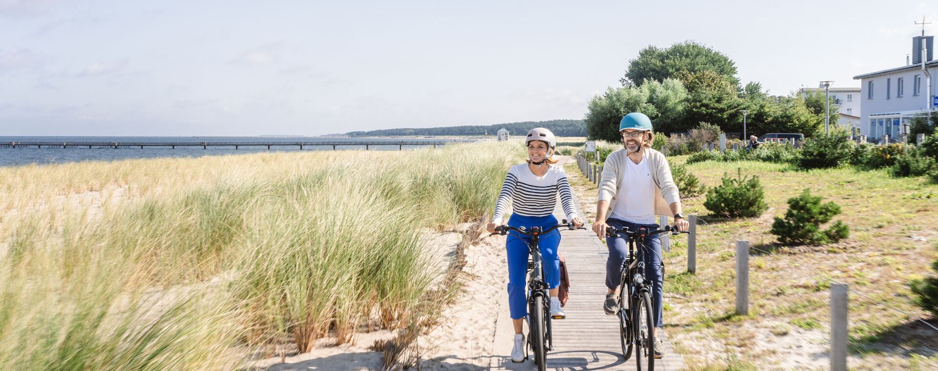 Zwei Fahrradfahrer auf einem Holzweg durch die Dünen. Links der Strand.