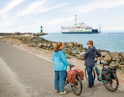 Zwei Frauen stehen mit dem Rücken zur Kamera und ihren Fahrrädern auf der Mole in Warnemünde. Auf der Ostsee fährt eine Fähre und im Hintergrund sieht man den grünen Leuchtturm