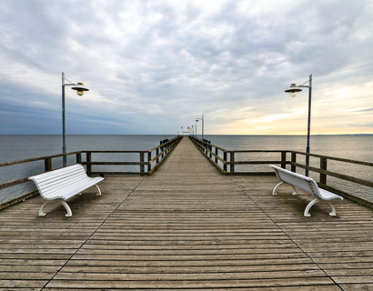 Das Bild wurde auf der Seebrücke stehend aufgenommen und zeigt die gesamte Länge der Brücke. Links und rechts stehen zwei weiße Bänke, die Sonne geht langsam unter und die Brücke ist umgeben von der Ostsee