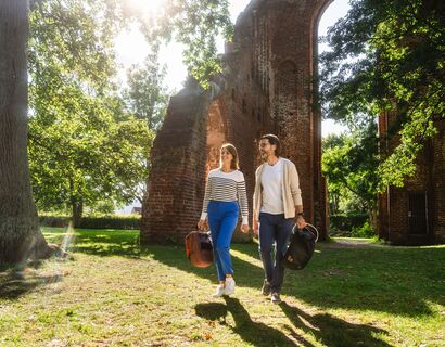 Paar spaziert im Schatten der Bäume. Im Hintergrund sieht man die Klosterruine.
