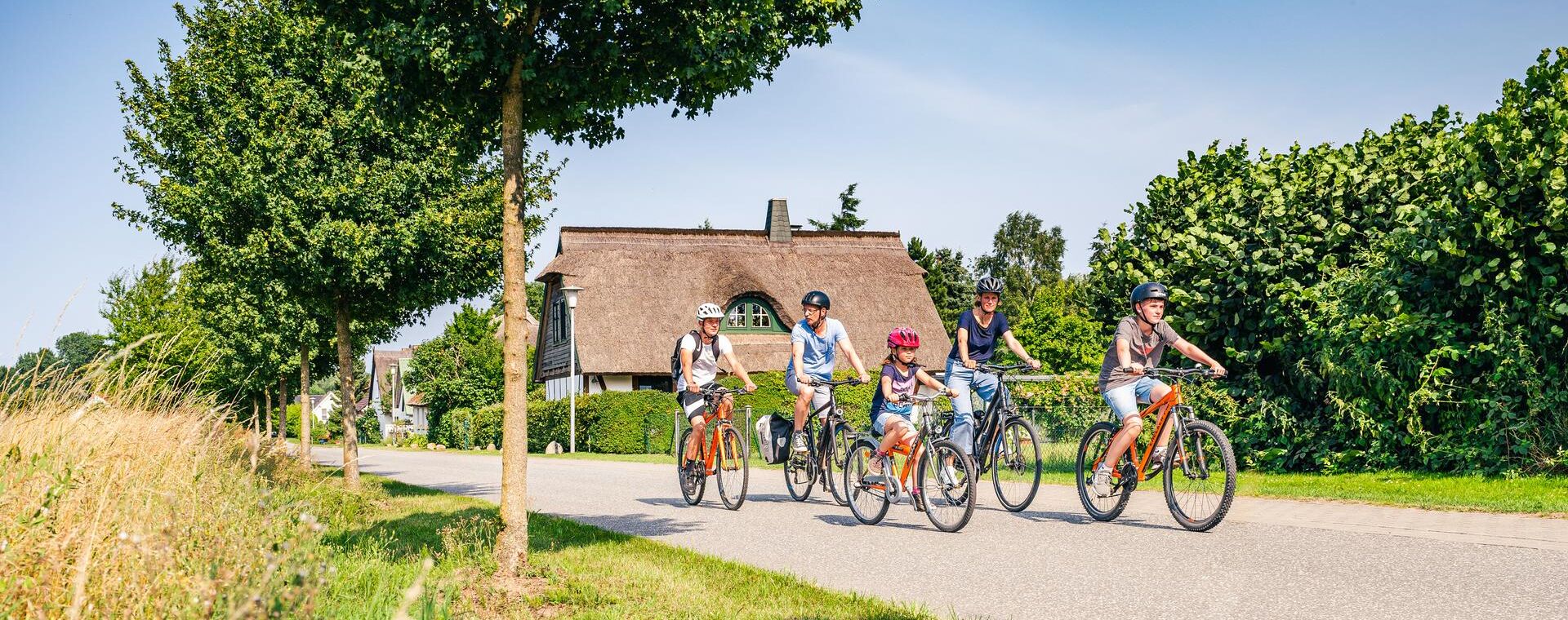 Eine Familie aus 5 Personen fährt Fahrrad. im Vordergrund ist ein begrünter Seitenstreifen und im Hintergrund ein Reetdachhaus