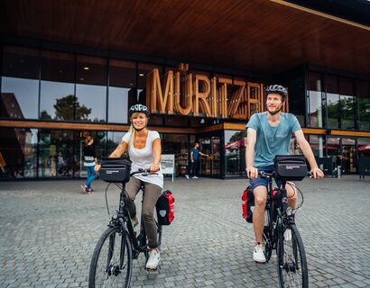 Ein paar startet seine Fahrrdtour vom Müritzeum aus. Beide schauen lächelnd in die Kamera, im Hintergrund sieht man den Eingang vom Müritzeum