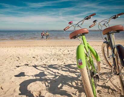 Rechts im Bild stehen ein grünes und braunes Fahrrad im Sand vom Strand. Im Hintergrund laufen zwei Personen durchs Wasser.