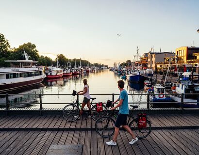 Zwei Radfahrer schieben ihr Rad über eine Brücke in Warnemünde in der Abenddämmerung und blicken aufs Wasser. Auf dem Wasser sind viele Boote.