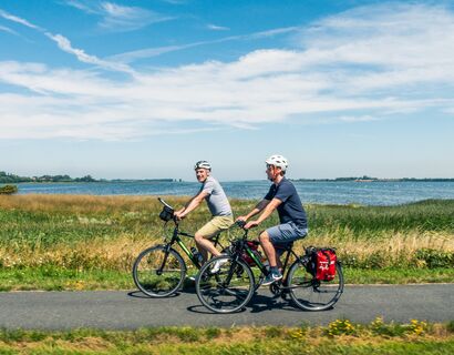 Zwei Männer fahren mit ihren Fahrrädern über einen aspahltierten Weg entlang von Wiesen und Gewässer. Die Radfahrer befinden sich mittig im Bild, einer der Männer schaut lächelnd in die Kamera.