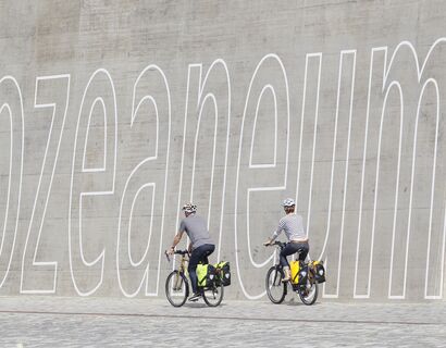 Zwei Radfahrer mit dem Rücken zur Kamera fahren mit ihren Fahrrädern an einer Wand vorbei. Auf der Wand steht in großer weißer Schrift "OZEANEUM"