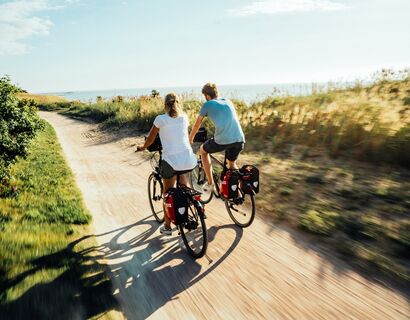 Zwei Fahrradfahrer fahren auf einem Sandweg entlang der Küste. Man sieht die Fahrradfahrer nur von hinten