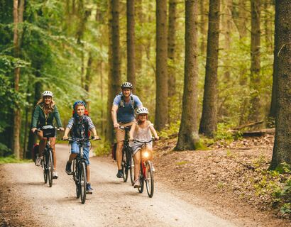 Eine Familie mit Eltern und zwei Kindern fahren mit ihren Fahrrädern durch einen Wald. Sie fahren auf die Kamera zu und lächeln