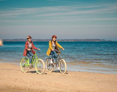 Zwei Fahradfahrerinnern, auf einem grünen und einem braunen Fahrrad über den Strand. Im Hintergrund das Wasser.