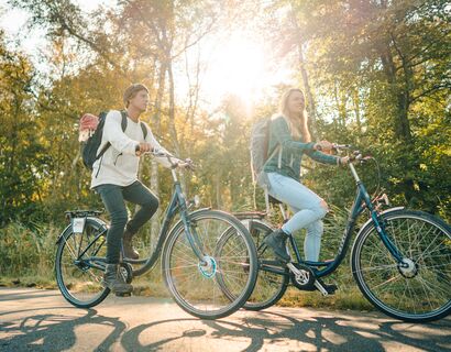 Ein Mann und eine Frau fahren auf Fahrrädern auf einem Asphaltweg durch den Wald. Das Bild wurde von unten aufgenommen, die Radfahrer sind im Fokus. Durch die beiden scheint hell die Sonne
